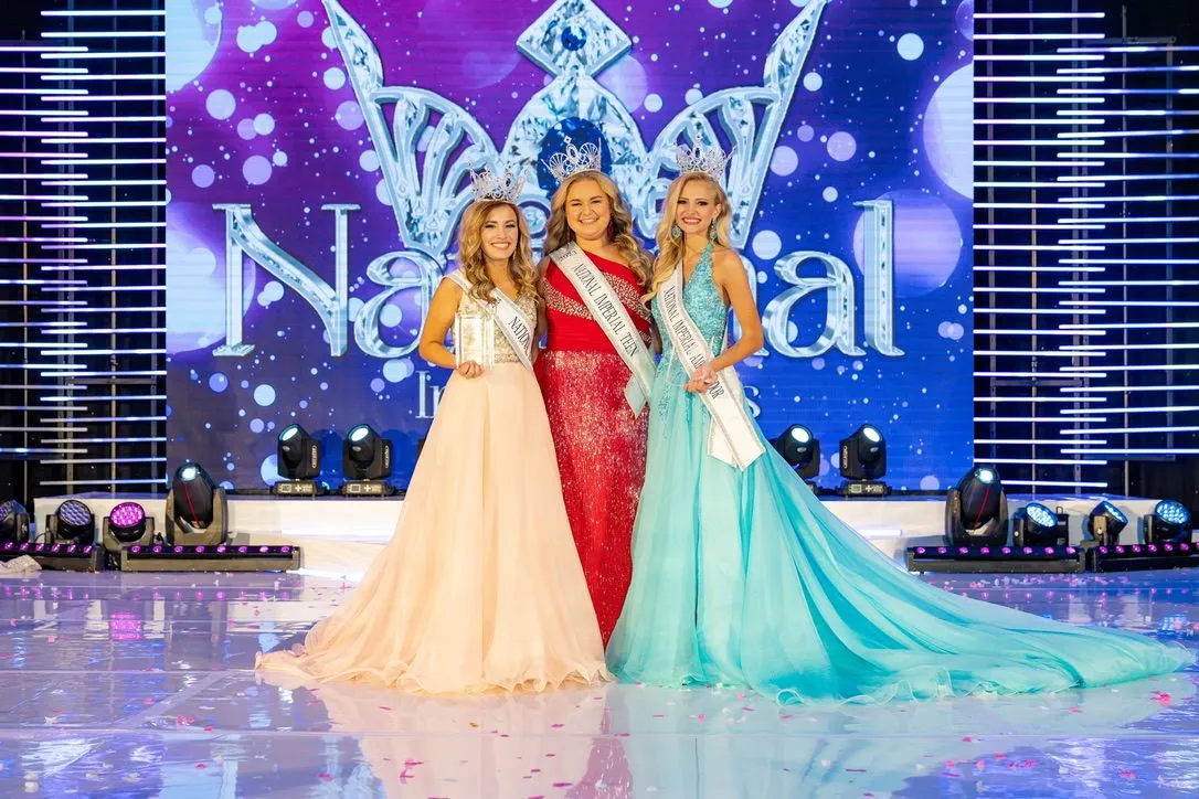 Three crowned titleholders in gowns on a national stage under a crown backdrop